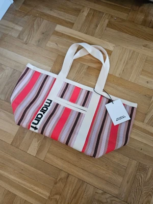 Randig shoppingväska Isabel Marant - Snygg shoppingväska från Isabel Marant i tyg med ränder i rosa, rött, beige och brunt. Väskan har vita handtag, en utvändig ficka och dragkedjeficka på insidan. Perfekt för att bära med sig allt man behöver.