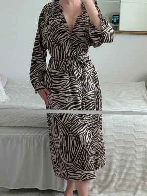Zebramönstrad satinklänning med bälte - Elegant zebra/mönstrad midi klänning i satinliknande material med knytning i midjan och flowy passform. Väldigt smickrande modell som passar perfekt till semester, middag, brunch eller sommar/fest 