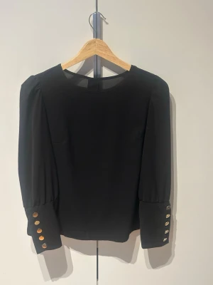 Svart blus med guldknappar - Stilren svart blus med rund halsringning och långa ärmar. Ärmsluten och ryggen pryds av dekorativast guldknappar. Blusen har en rak passform och är tillverkad i ett mjukt, lätt tyg som faller snyggt.