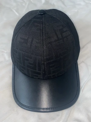 Svart Fendi keps - Snygg svart keps från Fendi med ton-i-ton FF-mönster på hela ovandelen och en böjd skärm i svart läder. Klassisk basebollmodell med exklusiv känsla och diskret logga. Perfekt accessoar för en trendig streetwear-look.