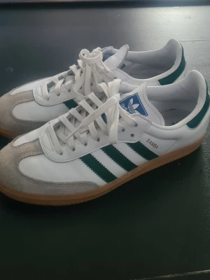 Adidas Samba vita och gröna sneakers - Klassiska Adidas Samba sneakers i vitt läder med gröna ränder och detaljer. Skorna har grå mocka framtill och gummisula. Ikonisk blå Adidas-logga på plösen och låga i modellen. Perfekta för en sportig och trendig stil.