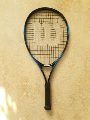 Wilson tennisracket svart/blå - Säljer ett Wilson tennisracket i svart och blått med stort W på strängarna. Racketen har svart grepp och är tillverkat i lätt och tåligt material, perfekt för dig som gillar att spela tennis och vill ha bra kontroll på banan.