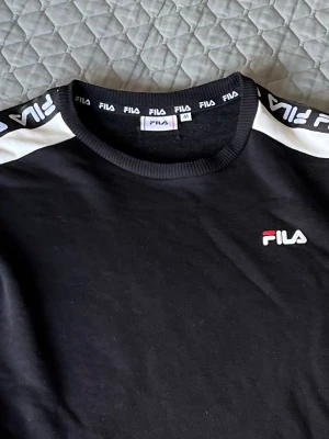Svart sweatshirt från FILA - Svart långärmad sweatshirt från FILA med vita detaljer och logga på bröstet. Tröjan har vita ränder med FILA-tryck längs ärmarna och ribbade muddar vid hals, ärmslut och nederkant. Perfekt för en sportig och avslappnad stil.