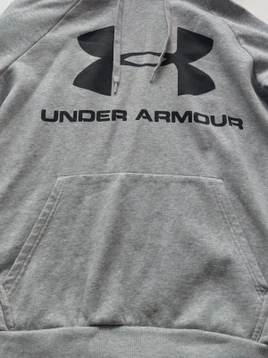 Grå hoodie från Under Armour - Snygg grå hoodie från Under Armour med stor svart logga och text på bröstet. Klassisk modell med huva och magficka. Perfekt för en sportig och avslappnad stil. Mjuk och bekväm bomullsblandning. Står storlek LG men skulle säga att den är mellan medium och large 
