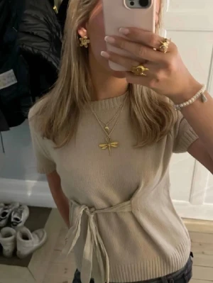 Beige stickad topp med knytband - Trendig beige stickad topp med korta ärmar och ett snyggt knytband i midjan. Toppen har rund halsringning och en mjuk, ribbad struktur som ger en stilren look. Perfekt att matcha med jeans eller kjol för en enkel men chic outfit. Pris kan diskuteras💗