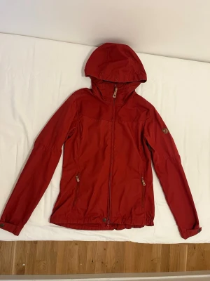 Röd vindjacka från Fjällräven - Snygg röd vindjacka från Fjällräven med huva och dragkedja framtill. Jackan har två fickor med dragkedja, justerbara ärmslut och en liten logotyp på ärmen. Tillverkad i slitstarkt material, perfekt för blåsiga dagar.