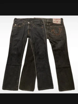 Svarta True Religion jeans bootcut - Säljer ett par svarta jeans från True Religion med klassiska kontrastsömmar och tydliga broderade bakfickor. Modellen har bootcut och låg midja. Jeansen är i denim och har fem fickor samt knappgylf.