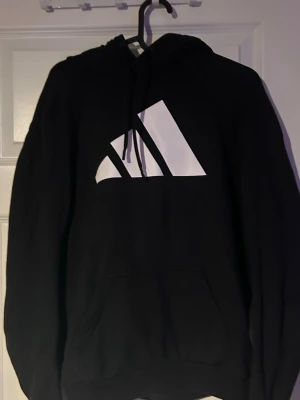 Svart hoodie från Adidas med logga - Svart hoodie från Adidas med stor vit logga på bröstet. Klassisk modell med huva
