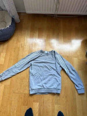 Grå Adidas sweatshirt med ränder - Snygg grå sweatshirt från Adidas med klassiska vita ränder längs ärmarna och diskret logga på bröstet. Tröjan har rund halsringning och är tillverkad i mjukt material, perfekt för en avslappnad streetstyle-look.