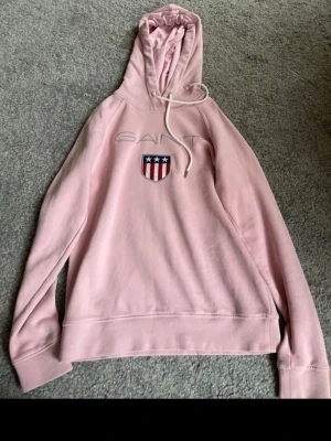 Ljusrosa hoodie från Gant - Säljer en ljusrosa hoodie från Gant med broderad logga och sköld på bröstet. Hoodien har dragsko i huvan och är tillverkad i mjukt bomullsmaterial. Perfekt för en avslappnad och snygg stil. Xs men väldigt stor i storlek så skulle säga mer s 