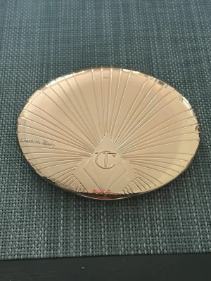 Charlotte Tilbury bronzer i guldig dosa - Snygg bronzer från Charlotte Tilbury i en lyxig, guldig snäckformad dosa med spegel. Pudret har en varm brun nyans som ger en solkysst look och är perfekt för att skulptera ansiktet. Dosan är i metall och känns riktigt exklusiv. Den är knappt använd men inte helt ny