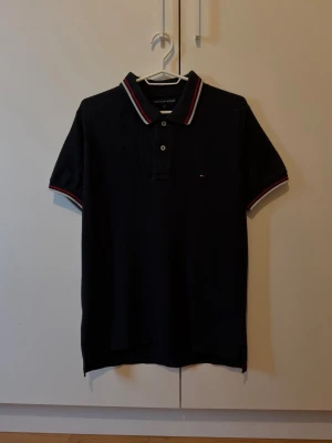 Svart piké från Tommy Hilfiger - Klassisk svart pikétröja från Tommy Hilfiger med vit och röd rand på krage och ärmslut. Tröjan har korta ärmar, knappar framtill och liten logga på bröstet. Perfekt till en clean och stilren look.