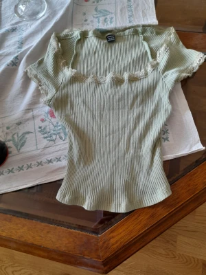 Ljusgrön ribbad topp med spets - Supersöt ljusgrön ribbad topp med korta ärmar och fina spetsdetaljer längs halsringningen och ärmsluten. Figurnära passform och stretchigt material som ger en bekväm känsla. Perfekt att styla med jeans eller kjol.