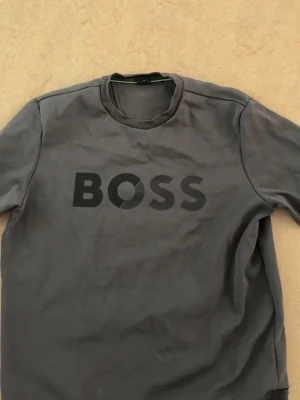 Grå BOSS t-shirt med tryck - Snygg grå t-shirt från BOSS med stort svart logotryck på bröstet. Klassisk rund halsringning och normal passform. Perfekt för dig som gillar stilrena och enkla plagg med en tydlig logga.