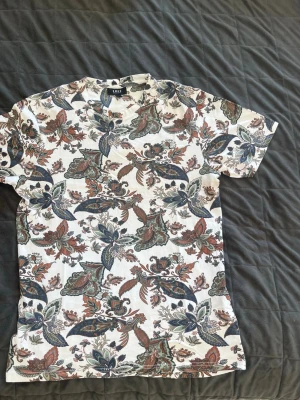 Vit blommig t-shirt från NEW YORKER - Snygg vit t-shirt från M&S med färgglatt blommigt mönster i grönt, blått, rött och brunt. Klassisk passform med rund hals och korta ärmar. Perfekt för dig som vill sticka ut med en unik och trendig stil.