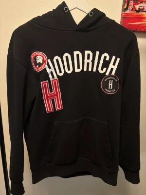 Svart hoodie från Hoodrich med tryck - Svart hoodie från Hoodrich med stora vita bokstäver och röda detaljer framtill. Har broderade patches, känguruficka och huva. Snygg vit signatur på axeln. Perfekt för streetwear-stil och chill dagar. Passar även som S, finns även byxor till vid intresse!!