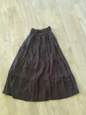 Svart lång kjol från H&M - Svart lång kjol från H&M med bred resår i midjan och flera volanglager. Kjolen är gjord i ett mjukt och lätt material, huvudsakligen viskos och polyamid, vilket ger ett snyggt fall och en avslappnad look.