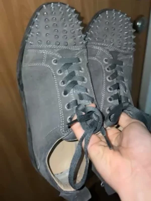 Gråa spikesneakers från Louboutin - Unika grå sneakers från Christian Louboutin med coola nitar över hela tån och klassiska röda sulor. Skorna är i mocka med svarta skosnören och vita kontrastsömmar. Perfekta för dig som vill sticka ut med din stil.