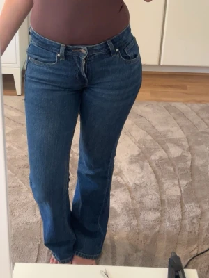 Blå bootcut jeans med låg midja - Säljer ett par klassiska blå bootcut jeans med låg midja och fem fickor. Jeansen har en snygg tvätt och raka ben som går ut lite nedtill. Perfekta till sneakers eller boots och ger en tidlös look.