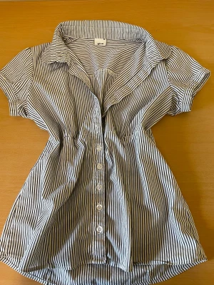 Randig kortärmad skjorta från Gina Tricot - Snygg skjorta från Gina Tricot med smala svarta och vita ränder. Skjortan har korta ärmar, klassisk krage och knappar framtill. Figursydd modell som ger en feminin siluett. Perfekt för en trendig och avslappnad look.