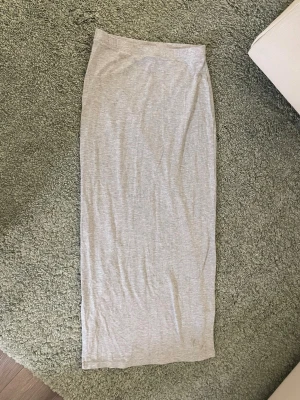 Ljusgrå långkjol med slits - En enkel och stilren ljusgrå långkjol i mjukt material. Kjolen har en rak passform och en diskret slits på sidan som ger extra rörelsefrihet. Perfekt att matcha med en crop top eller hoodie för en chill look.