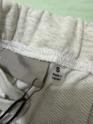Essentials Fear of God shorts ljusgrå - Ljusgrå shorts från Essentials Fear of God med elastisk midja och snörning. Klassisk och avslappnad passform, tillverkade i mjuk bomull. Logga tryckt på vänster ben. Perfekta för en chill och stilren look.