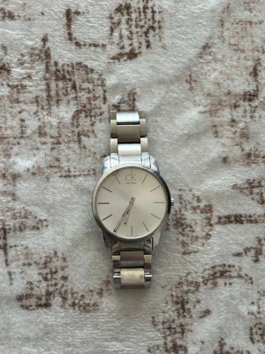 Silverfärgad klocka från Calvin Klein - Stilren klocka från Calvin Klein med rund, ljus urtavla och minimalistiska svarta markeringar. Armbandet är i blankt silverfärgat metall och har en klassisk länkdesign. Perfekt accessoar för dig som gillar enkel och modern stil. Klockan har en liten spricka i glaset, något som man kan fixa för en billig peng, annars är den i bra skick. Behöver ba lite kärlek o omsorg.