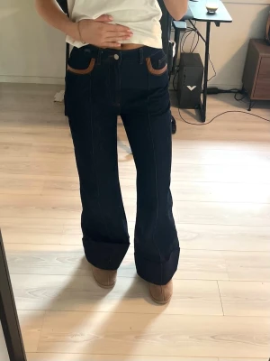 Mörkblå utsvängda jeans med kontrastfickor - Säljer ett par mörkblå jeans med utsvängda ben och bruna kontrastfickor. Jeansen är mid waist, synliga sömmar framtill och klassisk femficksmodell. Perfekta för dig som gillar retrovibbar och vill sticka ut lite extra. Dem är aldrig använda💕