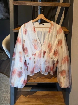 Blommig blus med v-ringning från Gina Tricot  - Säljer en superfin blus från Gina Tricot i glamsigt vitt tyg med stora rosa och röda blommor. Blusen har v-ringning, voluminösa långärmar och en lös, flowy passform. Perfekt för dig som vill ha en romantisk och trendig look.