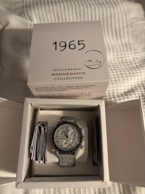 Omega x Swatch MoonSwatch klocka 1965  - Säljer en Omega x Swatch MoonSwatch 1965 från Bioceramic-kollektionen. Klockan har en ljusgrå textilrem med vita detaljer och logotyper, samt en vit urtavla med svarta och grå inslag. Stilig och unik design inspirerad av Speedmaster. Väldigt bra skick sparsamt använd hör av er vid frågor 👍