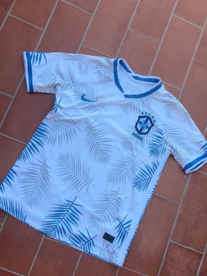 Vit, ljusblå Brasilien fotbollströja Nike S - Snygg vit och blå  fotbollströja från Nike med Brasilien-emblem på bröstet. Tröjan har blå detaljer vid krage och ärmslut samt ett mönster av palmblad i blått och grått över hela plagget. Perfekt för dig som älskar fotboll och vill sticka ut på planen. Sitter som S