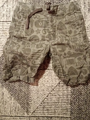 Gröna shorts med mönster och snörning - Säljer ett par gröna shorts med coolt mönster i olika former och figurer. Shortsen har snörning i midjan och flera fickor både fram och bak. Perfekta för sommaren när du vill ha en avslappnad och unik stil.