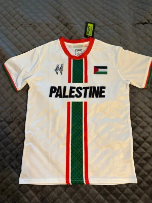 Vit Palestina fotbollströja  - Säljer en vit fotbollströja från PC Palestina med röd krage, gröna och röda ränder samt Palestina-flagga på bröstet. Tröjan har texten 'PALESTINE' i svart och detaljer i grönt och rött längs mitten. Perfekt för dig som vill visa ditt stöd på matchen.
