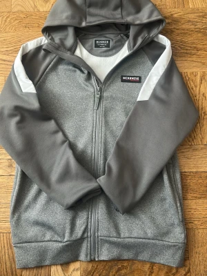 Grå hoodie från McKenzie med dragkedja 12-13 år - Snygg grå hoodie från McKenzie med hel dragkedja och huva. Tröjan har vita detaljer på axlarna och mjukt fleecefoder på insidan. Perfekt för en avslappnad stil och enkel att matcha med jeans eller joggers.