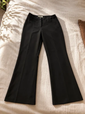 Svarta utsvängda kostymbyxor - Snygga svarta kostymbyxor med utsvängda ben och hög midja. Klassisk design med knapp och dragkedja framtill. Perfekta för en stilren look. Materialet känns slätt och följsamt.Inget fel me dom är de enda än ben som ni kan tabort om längd passar inte er