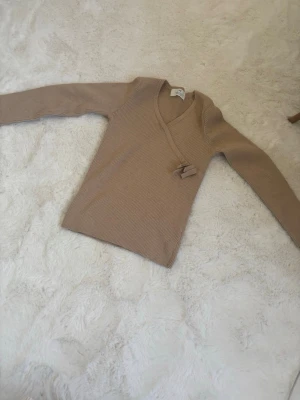 Beige omlottstickad tröja - Säljer en beige stickad tröja med omlottmodell och knytband i sidan. Tröjan har lång ärm och är ribbstickad vilket ger en snygg och smickrande passform. Perfekt att matcha med jeans eller kjol för en enkel men stilren look.