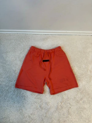 Röda Essentials shorts med snörning - Säljer ett par röda shorts från Fear of God Essentials med broderad logga på benet och snörning i midjan. Shortsen har en relaxed passform, elastisk midja och sidofickor. Perfekta för sommaren och riktigt sköna att bära.