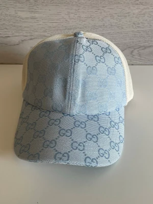 Ljusblå Gucci keps med GG-mönster - Säljer en snygg ljusblå keps från Gucci med klassiskt GG-mönster över hela framsidan och skärmen. Baksidan är i vit mesh för extra ventilation. Perfekt för dig som vill ha en trendig och exklusiv accessoar till din outfit.