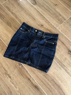 Mörkblå jeanskjol med fickor - Snygg mörkblå jeanskjol från Cubus med klassisk femficksdesign och synliga sömmar. Kjolen har bälteshällor och knappstängning framtill. Perfekt för dig som gillar en enkel och tidlös stil.