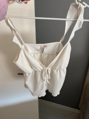 Vit linnetopp med knyt från H&M - Säljer en vit linnetopp från Nelly med v-ringning och knyt framtill. Toppen har smala axelband och är tillverkad i ett lätt, strukturerat bomullsmaterial. Perfekt för varma dagar och enkel att matcha med jeans eller kjol.