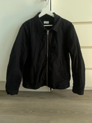 Svart jacka från Tiger of Sweden - Bara lite urtvättad men inte mycket. Nypris nära 3000kr. Stilren svart jacka från Tiger of Sweden med klassisk krage och dragkedja framtill. Jackan har en enkel, clean look och är tillverkad i ett mjukt bomullsmaterial. Perfekt för dig som gillar minimalistisk stil.