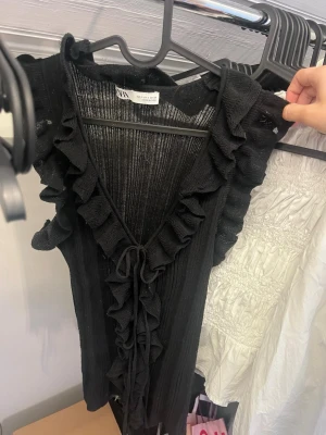 Svart ribbad topp med volang - Supersnygg svart ärmlös linnetopp från zara med ribbad struktur och volangdetaljer längs v-ringningen. Toppen har knyt framtill och är perfekt för att lyfta vilken outfit som helst. Lätt och luftig känsla, passar perfekt till jeans eller kjol.