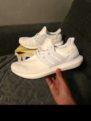 Adidas Ultra Boost vita sneakers - Fräscha helvita Adidas Ultra Boost sneakers med stickad ovandel och klassiska tre ränder på sidan. Skorna har en chunky Boost-sula för maxad komfort och sportig vibe. Perfekta för dig som gillar stilrena och cleana sneakers med modern känsla.