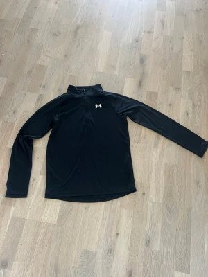 Svart träningströja från Under Armour, stl Junior L - Svart långärmad träningströja från Under Armour med halv dragkedja och hög krage. Tillverkad i ett stretchigt och funktionellt material som andas. . Diskret logga på bröstet. Perfekt på golfbanans eller på gymmet⛳️🏌🏼‍♂️💪! 