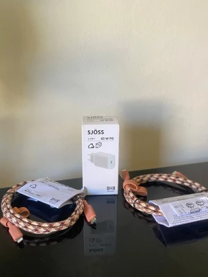 SJÖSS 65W PD laddare med flätade kablar - Säljer en SJÖSS 65W PD USB-C laddare i vitt tillsammans med två flätade USB-C till USB-C kablar i beige, brunt och rött mönster. Kablarna har gummiliknande kontakter i ljusrosa och är tåliga och flexibla. Perfekt för snabbladdning av mobil, surfplatta eller laptop.
