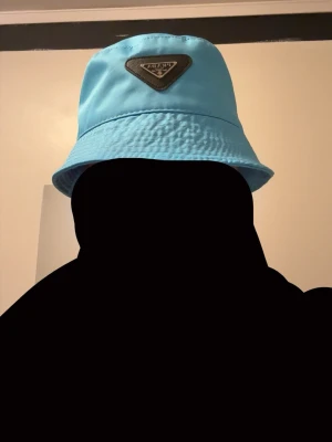 Blå bucket hat från Prada - Snygg ljusblå bucket hat från Prada med klassisk trekantig logotyp framtill. Hatten är tillverkad i ett lätt och bekvämt material som passar perfekt för en trendig streetstyle-look. Perfekt accessoar för att lyfta din outfit. Perfekt nu till sommaren!!