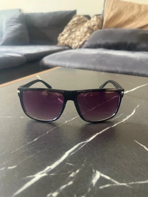 Svarta solglasögon med lila ton - Snygga solglasögon med svart båge och rektangulär form. Glasen har en mörk lila ton som ger en cool vibe. Perfekta för att lyfta din stil till nästa nivå. Klassisk design som passar till det mesta.