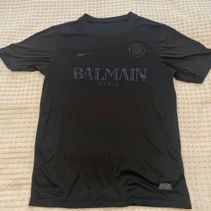 Svart PSG x Balmain t-shirt Nike - Snygg svart t-shirt från Nike i samarbete med PSG och Balmain. Tröjan har rund hals, korta ärmar och tryck med Balmain Paris-logga samt PSG-emblem på bröstet. Tillverkad i ett lätt och sportigt material, perfekt för träning eller streetwear.