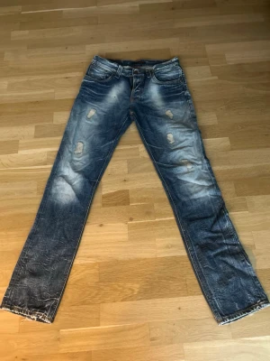 Blå slitna nudie jeans - Säljer ett par blå nudie jeans med raka ben och slitna detaljer framtill. Jeansen har klassisk femficksmodell, ljusa tvättade partier och råa kanter vid bensluten. Perfekta för en avslappnad och trendig look.