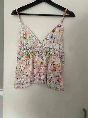 Blommig spetslinnetopp med broderi - Superfin linnetopp med blommigt mönster i pastellfärger och v-ringning. Toppen har tunna axelband, broderade detaljer och spetsinslag som ger en romantisk känsla. Perfekt för varma dagar och somriga outfits.